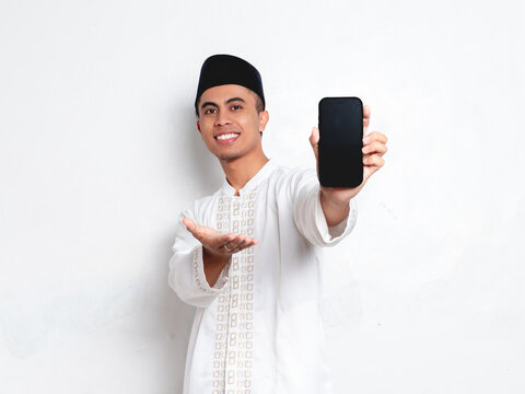 Excited Indonesian Muslim man showing blank smartphone screen for mockup, wearing peci and baju koko on white background, celebrating Ramadan sale promotion concept