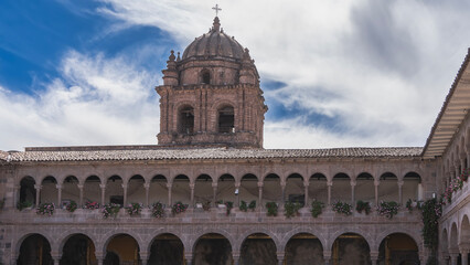 An ancient monastery Church and Convent of Santo Domingo. The courtyard. Red stone walls. Galleries with columns and arches. A bell tower with a dome and a cross against a blue sky, clouds. Peru.Cusco