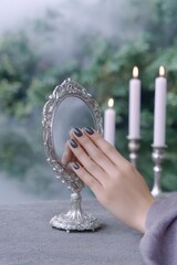 a witchs hand with black nails with antique mirror, surrounded by candles. the atmosphere is mystical, with a smoky fog in the background and dark gray and green tones