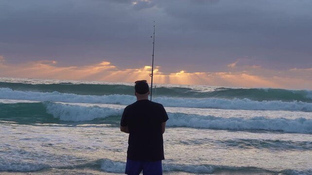 Rear view of a fisherman walking into rolling ocean waves at dawn holding a fishing rod, with strong sun rays streaming through the sky over the sea.