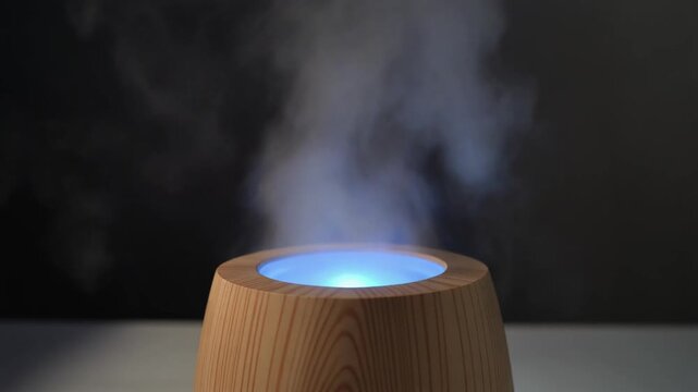 Humidifier with Wood-Grain Finish and Blue Illumination &mdash; A humidifier finished in wood grain is placed on a surface, releasing visible vapor from its top. It features a gentle blue glow surrounding i