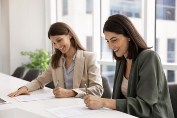 Two professional women sitting at office table, smiling and signing legal documents, put signatures on business agreement or contract. Successfully closing deal, formalizing new business collaboration