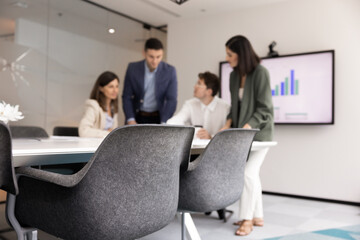 Four professionals, analysts gathered around conference table in office, engaged in collaborative discussion, digital screen behind them displaying bar charts. Presentation preparation or strategizing