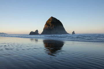 Haystack Rock at sunrise on Cannon Beach, Oregon Coast, USA with Pacific Ocean tides, wet sand reflections, and surrounding sea stacks in soft morning light. Ideal for travel, tourism, nature themes