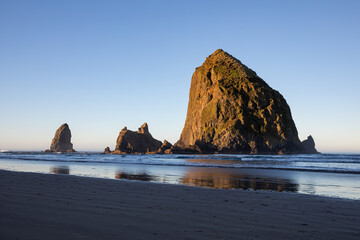 Haystack Rock at sunrise on Cannon Beach, Oregon Coast, USA with Pacific Ocean tides, wet sand reflections, and surrounding sea stacks in soft morning light. Ideal for travel, tourism, nature themes