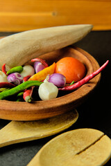 Assorted fresh ingredients including chilies, shallots, garlic, and tomato in a traditional wooden mortar with rustic kitchen tools, top view food still life.