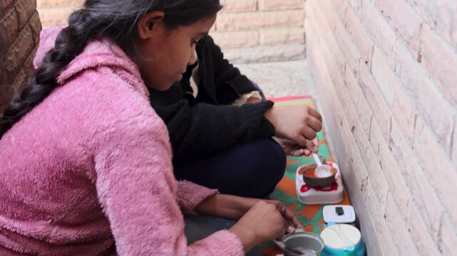 Two village girls playing pretend kitchen game with small utensils in a rural home. Childhood joy, imagination and simple village lifestyle in Uttarakhand, India.