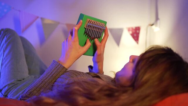 Young woman lying comfortably on bed, gently playing green kalimba, surrounded by festive flags and soft ambient lighting, creating peaceful musical moment