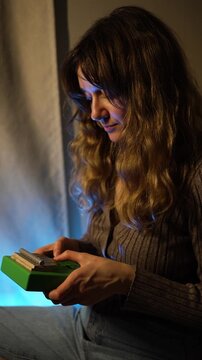 Young woman musician with long brown hair sits and plays a green kalimba illuminated by blue light in a dimly lit room, creating a peaceful and artistic atmosphere