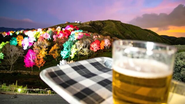 A refreshing glass of beer on a tray with a colorful illuminated forest in the background at dusk.