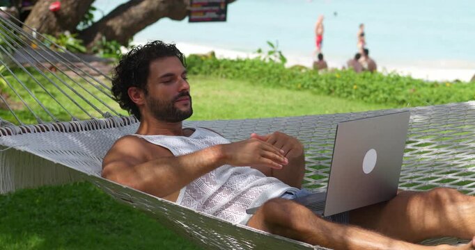 Relaxed young Caucasian male tourist on vacation resting in beach hammock against the beach, talking with friends via video call on laptop. Online meeting and communication from anywhere in the world