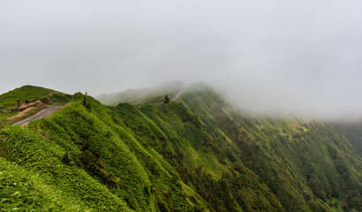 Naklejka premium Sete Cidades volcanic landscape in San Miguel, Azores. Lush green hillsides covered in fog and mist. Scenic view of the natural environment. Remote outdoor setting with winding path.