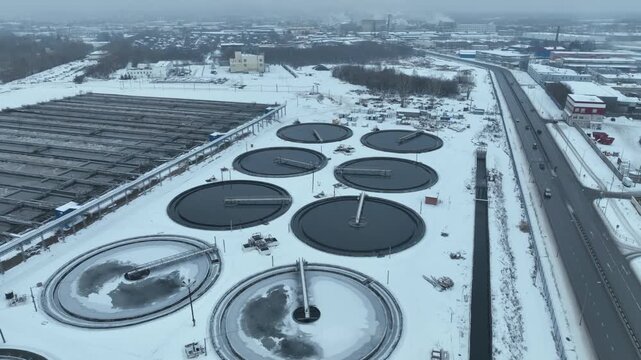 Frozen aeration basins display cracked icy surfaces. Harsh winter conditions intensify industrial austerity and technical severity