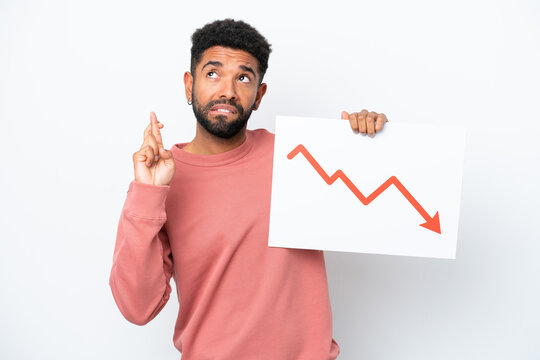 Young Brazilian man isolated on white background holding a sign with a decreasing statistics arrow symbol with fingers crossing