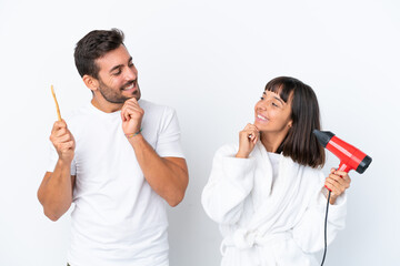 Young caucasian couple holding a hairdryer and toothbrush isolated on white background looking looking at each other