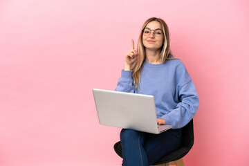 Young woman sitting on a chair with laptop over isolated pink background pointing with the index...