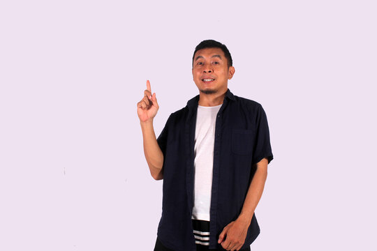 A happy looking Asian man wearing a blue shirt with a white undershirt points to an empty space, isolated on a white background.