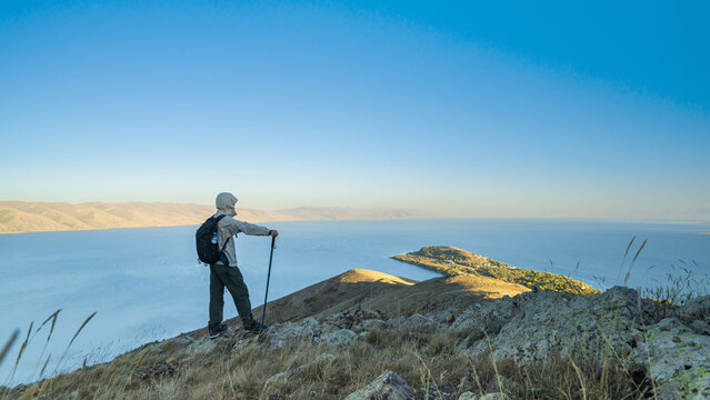Hiker with backpack standing on a rocky hill above Lake Sevan, Armenia, looking at the wide blue water and distant mountains under clear sky.