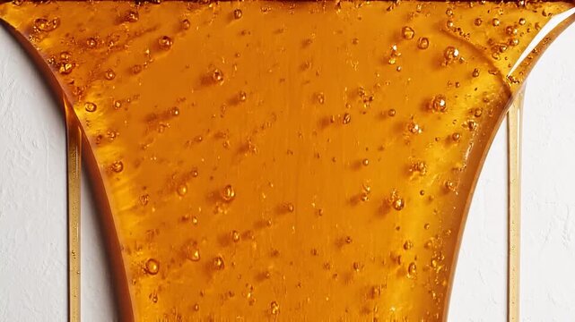 Luxurious golden honey with tiny reflective bubbles slowly drips down a clean, textured white surface, creating an appetizing and elegant abstract foo