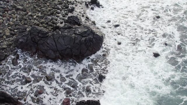 Cinematic drone shot moving backwards to reveal large dark volcanic boulders and white sea foam on a rocky tropical shoreline in East Nusa Tenggara.