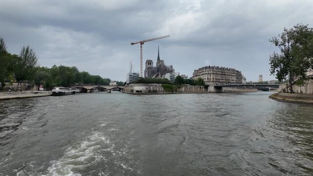 Navigating the seine river away from notre dame cathedral in paris, france during reconstruction under a cloudy sky with cranes and scaffolding on the famous landmark