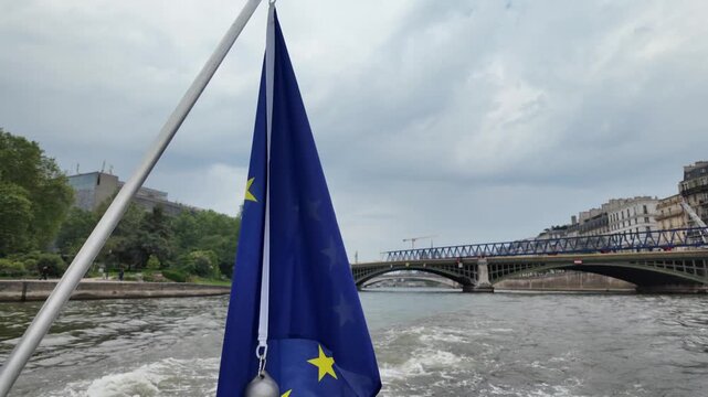 European union flag waving on a tour boat cruising along the seine river in paris, with a picturesque view of the pont d'austerlitz bridge and city buildings under a cloudy sky