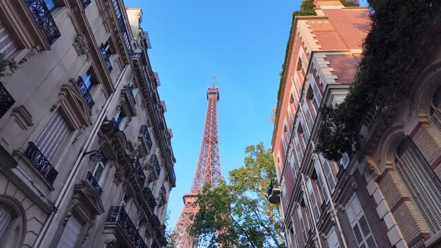 Revealing eiffel tower from a parisian street with traditional haussmann buildings and green trees against a clear blue sky, low angle dolly out shot in paris, france