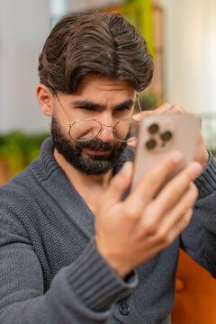 Indian confused man at home squints at smartphone with eyeglasses tries to read small text blinks adjusts frame feels strain a bit. Arabian guy on sofa holds phone near face uses zoom checks message