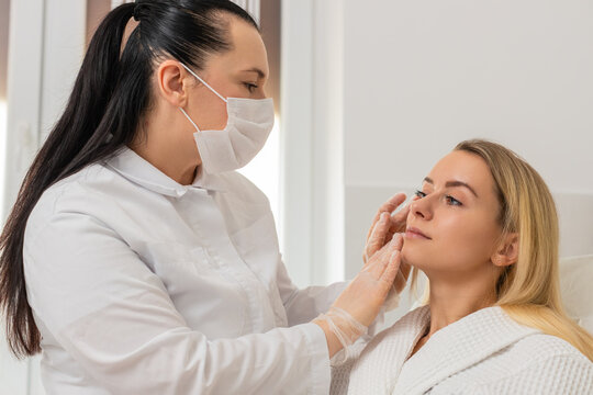 Young woman sits in chair while cosmetologist in mask gently inspects facial skin before treatment. Adult client in white robe feels calm trusts professional skincare evaluation in modern clinic room.