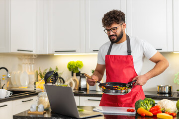 Arabian man cooks vegetables in frying pan while checking laptop recipe in kitchen for healthy meal...