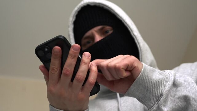 A scammer unlocks a phone to withdraw money.
A masked man holding a phone.
Withdrawing money online from a phone.