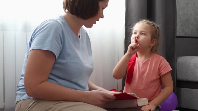 Mid shot of young Caucasian woman speech therapist reads book with cute girl. Mother and daughter learning together. Preschool education for young children.