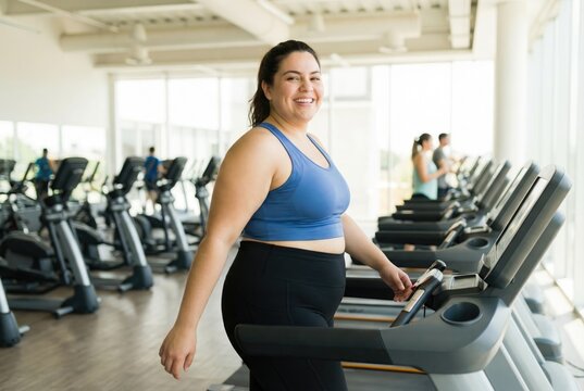 Happy overweight woman smiling while walking on a treadmill in a fitness center. Plus size girl enjoying a cardio workout for weight loss.