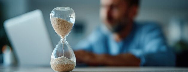 Hourglass in foreground frames focused worker using laptop behind blur.