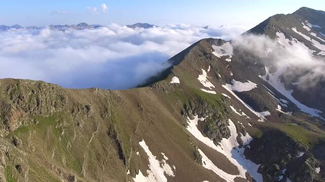 Aerial view of moist air condensing along a high altitude mountain ridge slope in the United States. Orographic cloud formation spreads across rugged alpine terrain under dynamic mountain weather.