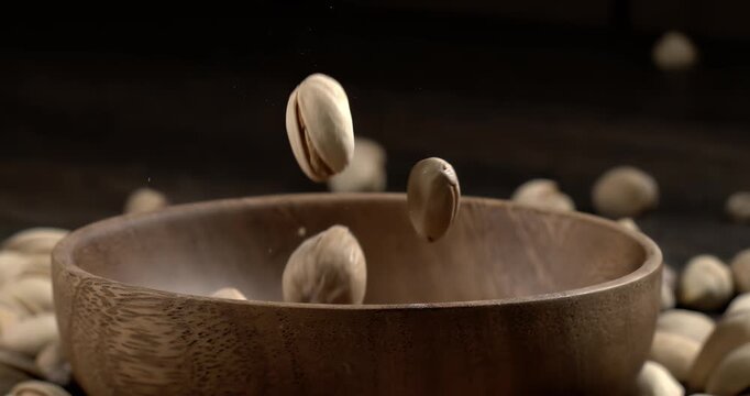 Wooden bowl filled with pistachios on dark wooden table, tasty salted nuts scattered around, healthy snack and natural food concept. Shot on super slow motion camera 1600 fps.