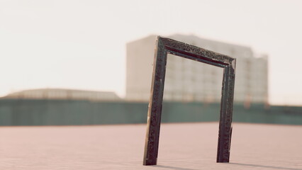 A unique frame stands upright on a sandy expanse, capturing the warm hues of a sunset. The scene is serene, with distant buildings softly blurred in the background. © icetray