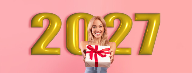 Obraz premium A woman stands smiling and holding a wrapped gift. Behind her are large numbers showing the year 2027. The background is pink. She appears happy and excited about the gift.