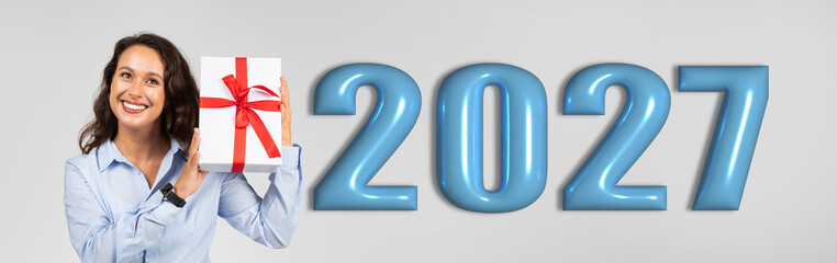 Obraz premium A woman smiles while holding a gift box decorated with a red ribbon. Large numbers for the year 2027 are displayed beside her. This scene suggests a celebration or special occasion.