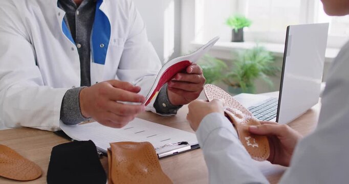 Hands of orthopedist show insoles to patient at consultation in podiatry clinic. Doctor explaining to woman layered orthotic insoles as arch support for foot pain, corrective flatfoot treatment