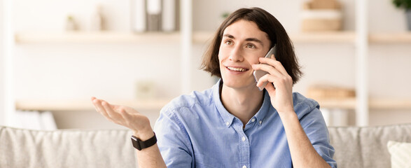 Fototapeta premium A man is sitting on a couch and talking on his smartphone with a cheerful expression. He gestures with one hand while engaged in the conversation. The setting is cozy and well-lit.