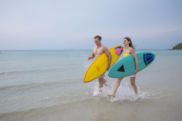 Obraz premium Happy couple running with surfboards in tropical sea, enjoying summer vacation and outdoor surfing lifestyle. Ideal for water sports, active travel, beach fun and fitness themes.