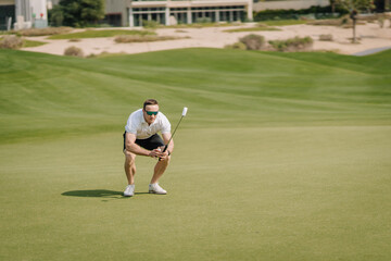 Male golfer crouching on green grass course preparing to putt with golf club, sunny day and manicured landscape visible in the background