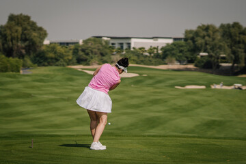 Female golfer in pink shirt and white skirt swings club on green golf course with manicured grass and trees in the background under clear sky