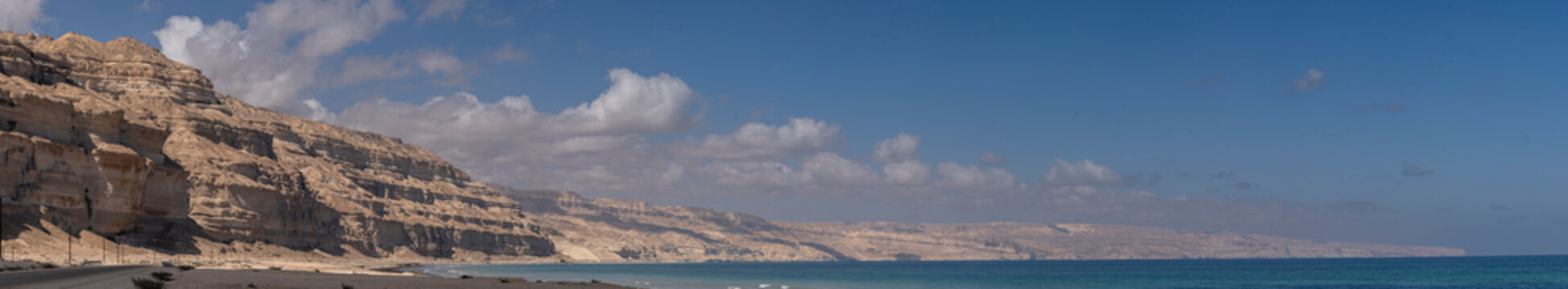 Wide coastal panorama along Mirbat&ndash;Hasik Road, Oman: rugged limestone cliffs meeting turquoise Arabian Sea under a clear blue sky with scattered clouds.