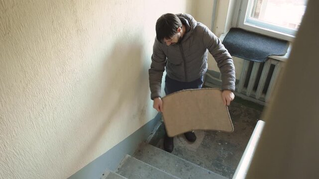 Caucasian man in jacket manually shakes dust out of rug on stairwell landing. Person performs household chores in authentic municipal house on back staircase.