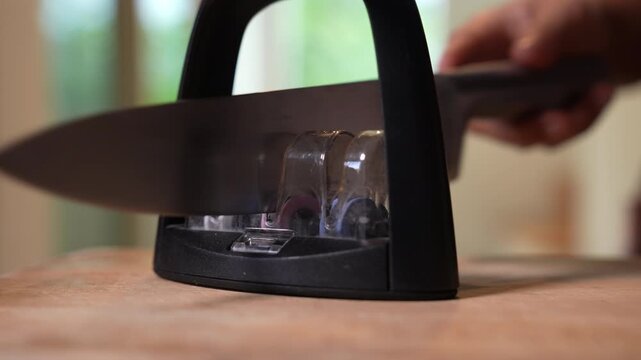 Close-up of kitchen knife being sharpened on a manual knife sharpener tool, hand sharpening blade in domestic kitchen, home cooking preparation and kitchenware maintenance.