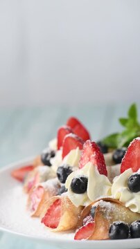 Thin crepes filled with fresh berries, topped with whipped cream, strawberries, and blueberries, dusted with powdered sugar and mint. Close-up of the dessert on a white plate. Vertical video. Circular
