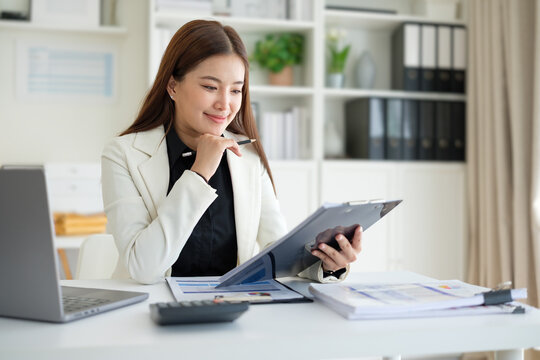 Focused professional businesswoman working on a laptop while reviewing documents on a clipboard.