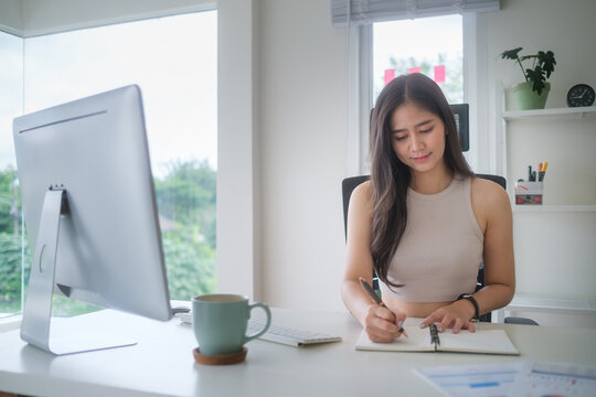 Young businesswoman writing notes in a notebook while working at her desk in a bright modern office with laptop and coffee mug.
